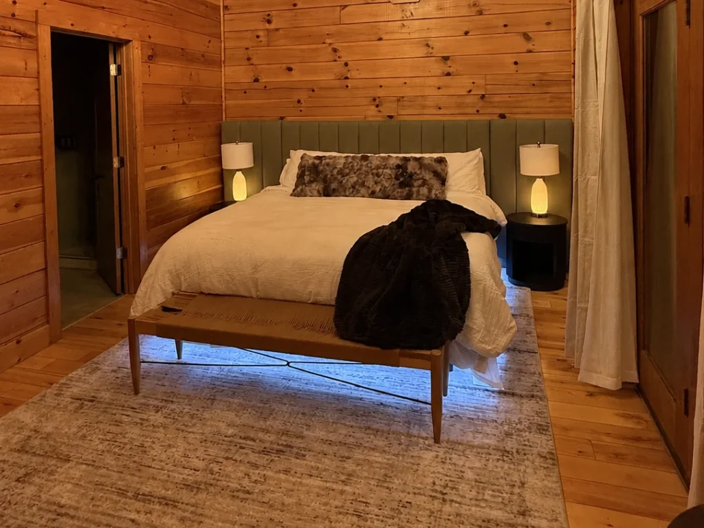 King bed with an olive upholstered headboard, dark throw and woven bench beside curtained deck doors.