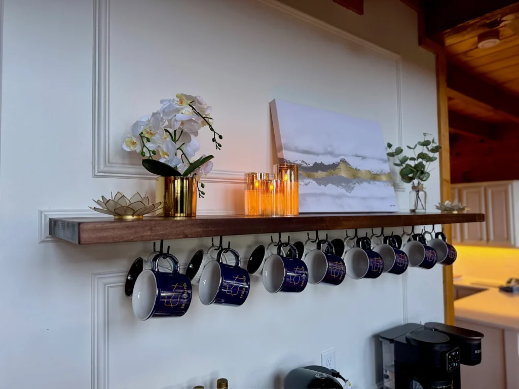 Mugs hang beneath a shelf of flowers, mountain art and warm accent lights.