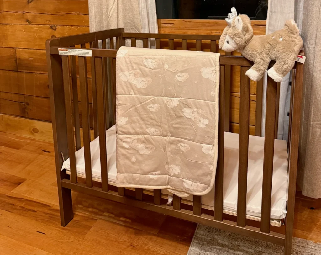 Wooden crib with a cream quilt and plush deer beside the cabin window.