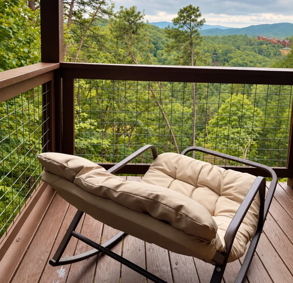 Cushioned rocking chair on a covered deck overlooking wooded mountains, with neighboring cabins visible on the far hillside.