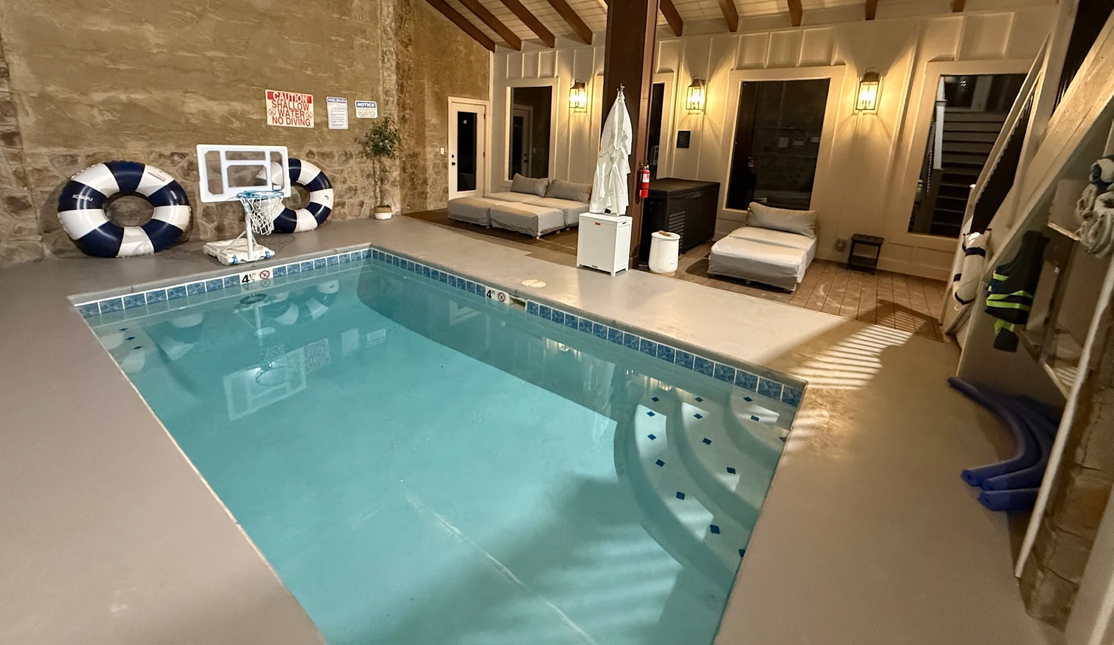 Indoor pool with curved submerged steps, white loungers, stone walls and warm room lighting.