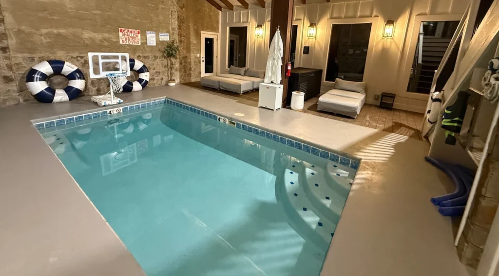 Indoor pool with curved submerged steps, white loungers, stone walls and warm room lighting.