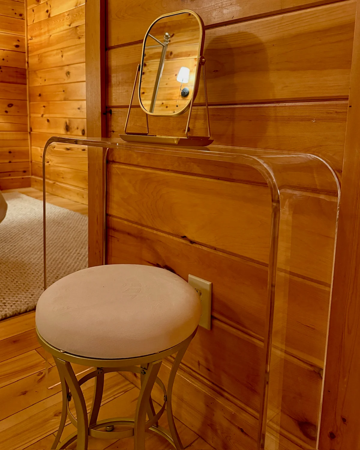A clear makeup vanity with a gold-framed mirror and a round upholstered stool against a wood wall.
