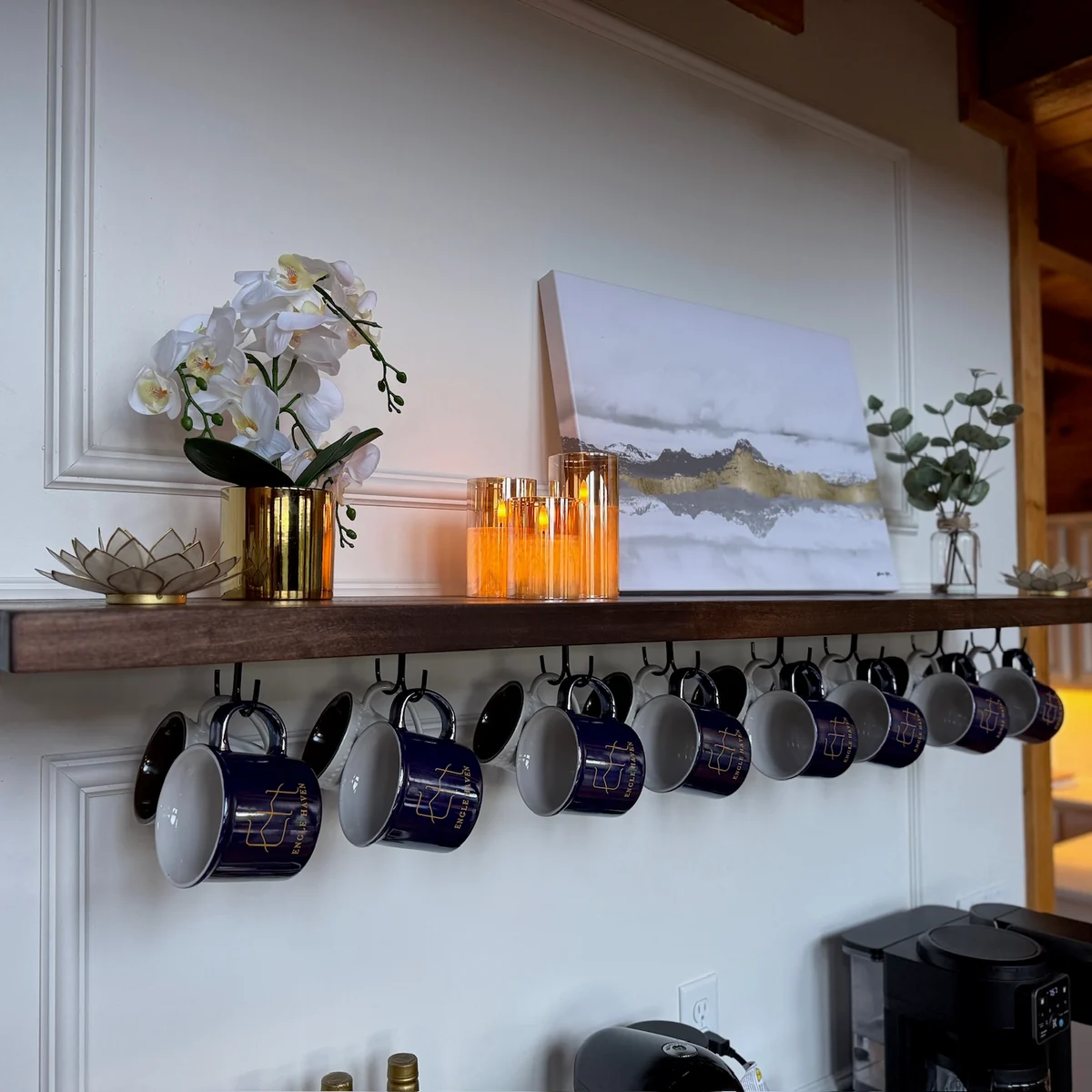 Flowers, landscape artwork and warm candlelight arranged on a shelf above hanging Engle Haven mugs.