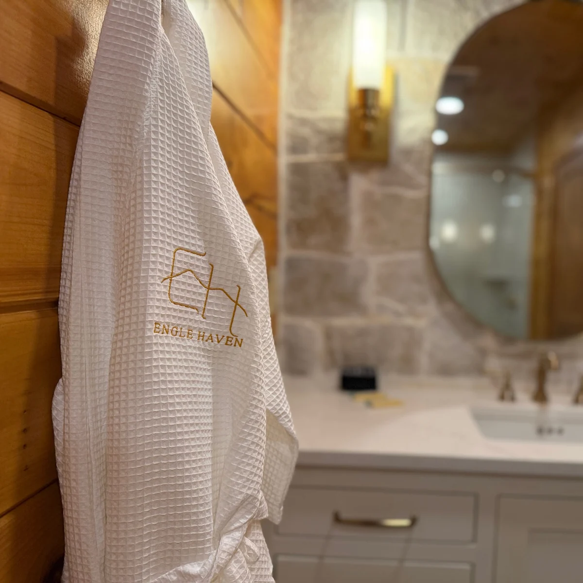 A white robe embroidered with the Engle Haven logo hangs beside a softly lit bathroom vanity.