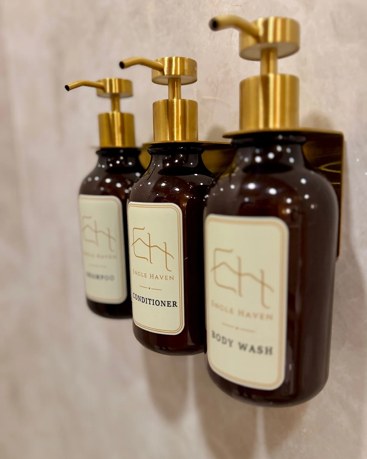 Three amber Engle Haven bath-product bottles with gold pumps mounted on a tiled wall.