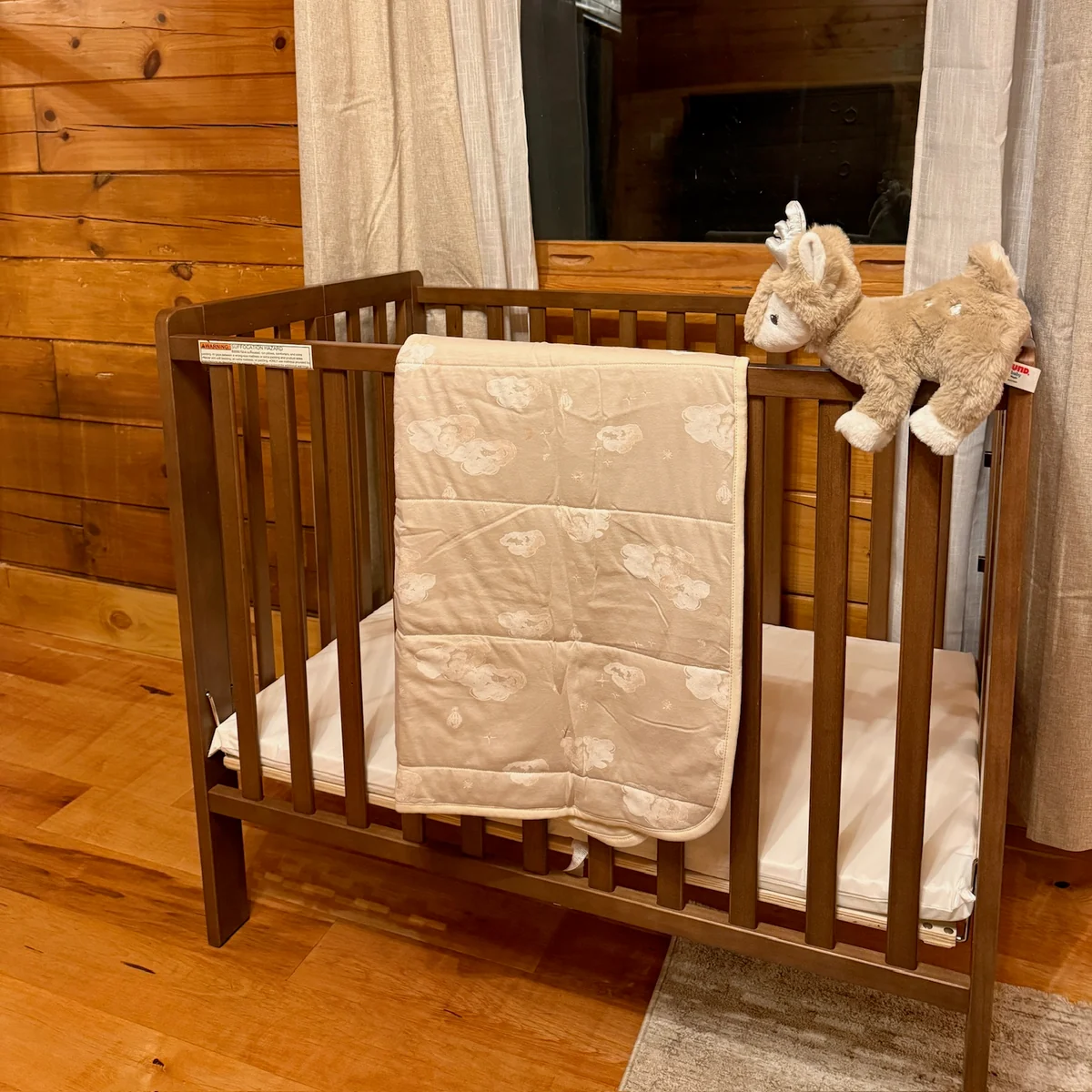 A wooden crib beneath a curtained window, with a light quilt draped over the front rail.