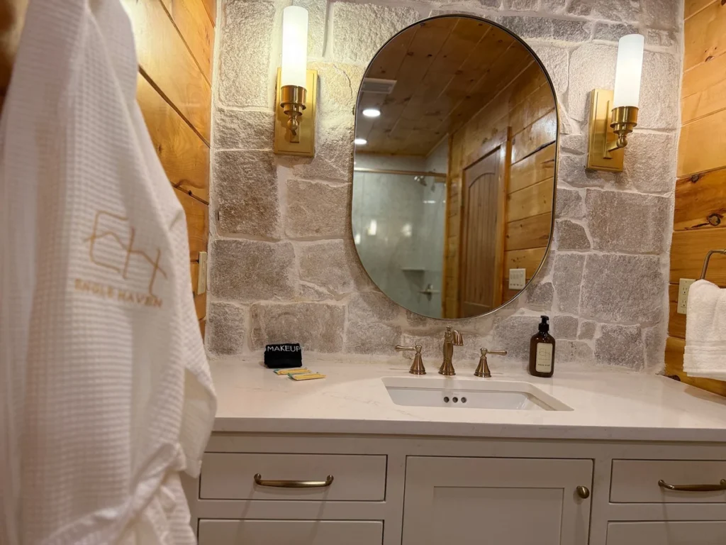 Branded white robe beside an oval bathroom mirror, brass lights and faucet, and a white vanity.