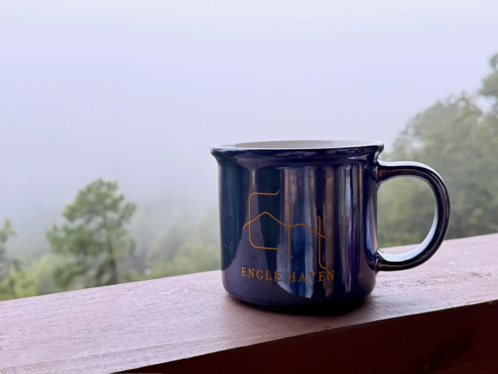 Engle Haven mug on a deck railing with mist over the wooded mountains.