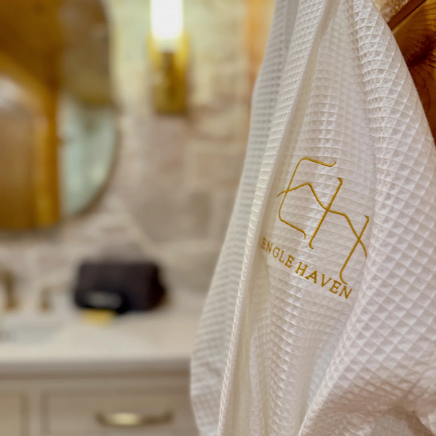 Gold Engle Haven embroidery on a white waffle-textured robe, with the bathroom softly blurred behind it.