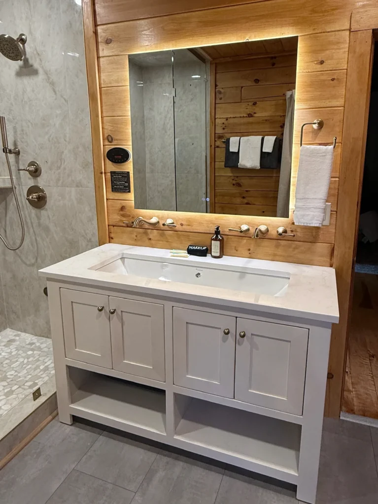 Illuminated mirror and white vanity beside the steam-shower fixtures and wall-mounted steam control.