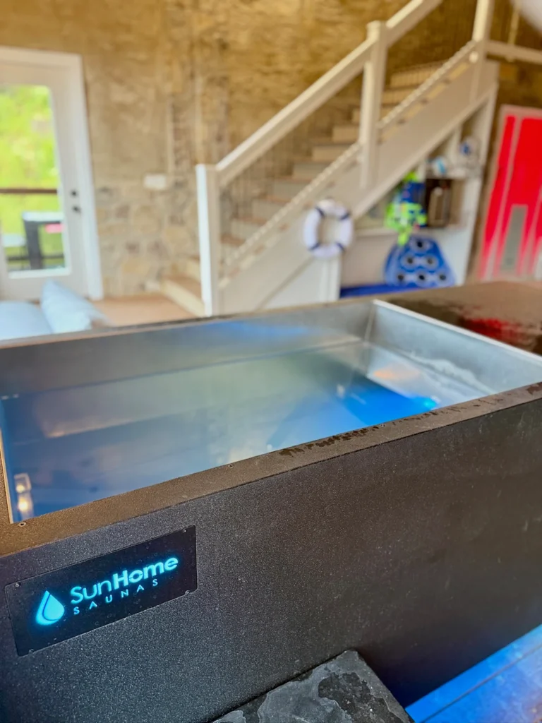 Water-filled cold-plunge tub, with the pool-room stairs and red-lit sauna behind it.