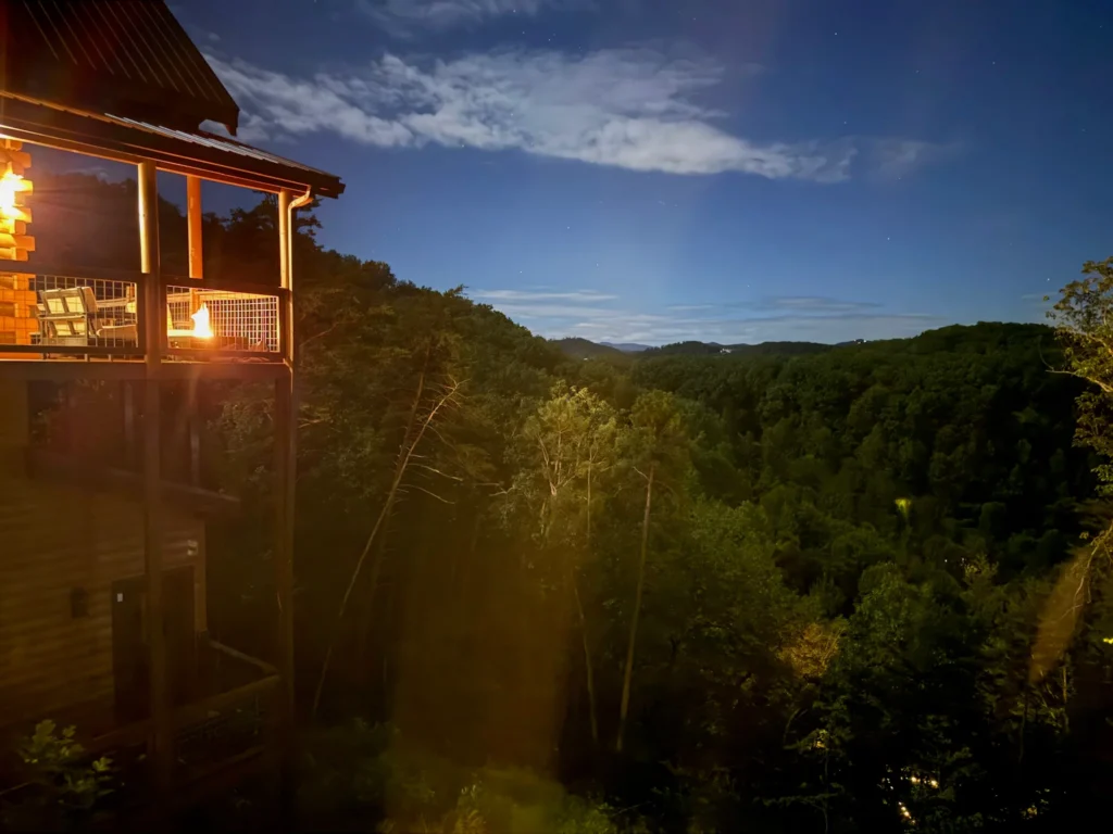 Lit cabin deck and fire table beside wooded hills at dusk.