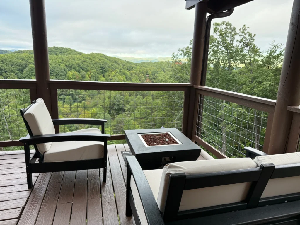 Two cushioned chairs and a square fire table on the covered deck overlooking wooded hills.