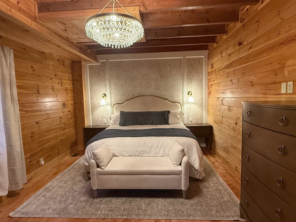 Upholstered king bed with a neatly laid dark runner, matching bench and crystal chandelier in a timber-lined bedroom.