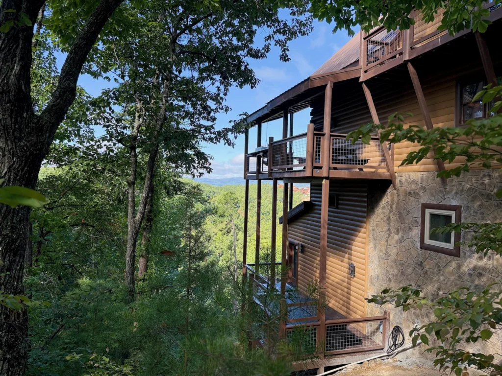 Engle Haven’s timber decks and stone lower level beside a wooded hillside, with the distant mountain setting visible between the trees.